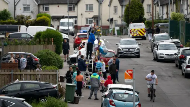 People climb a ladder to put flowers around a lamp-post to pay tribute to the two teens killed in a crash that sparked a riot.