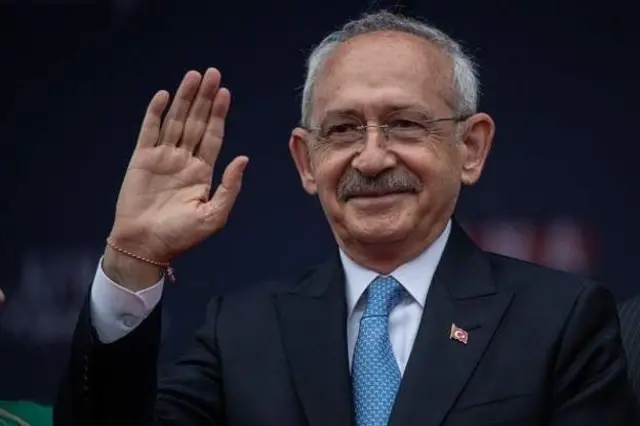 Kemal Kilicdaroglu, a man with short grey hair, a moustache and glasses, waving. He is wearing a suit with a blue tie and has string bracelet on his right hand.