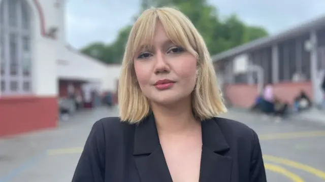 A picture of Husna Donmez. She is a young woman with blonde hair, wearing a black blazer. The background behind her is blurred