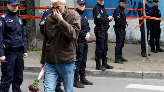 Man holding bouquet of flowers wipes tears away as he passes a line of police officers manning the scene.