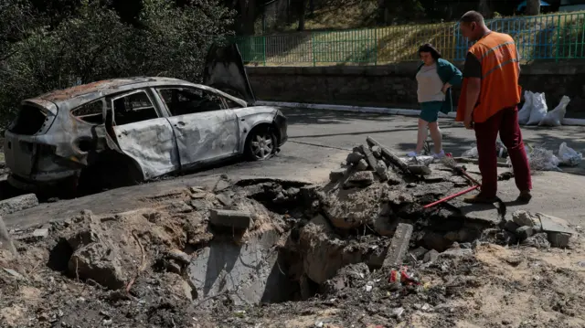 Two people stand near a shell hole after debris from a drone attack fell outside an apartment building in Kyiv