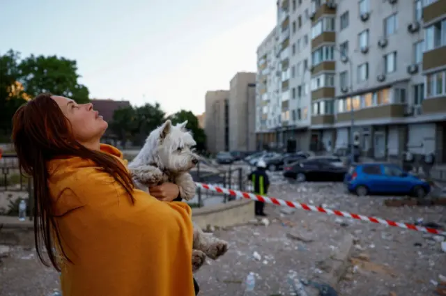 A woman with a dog looks at her heavily damaged apartment building