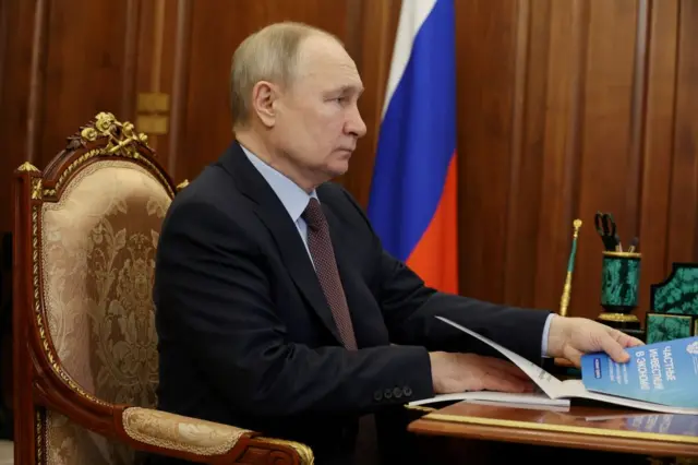 Vladimir Putin looks solemn as he sits in chair holding a notepad