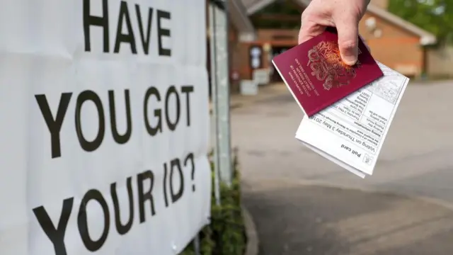 Stock image of a hand holding a passport and a voting card near a sign which says "have you got your ID?"