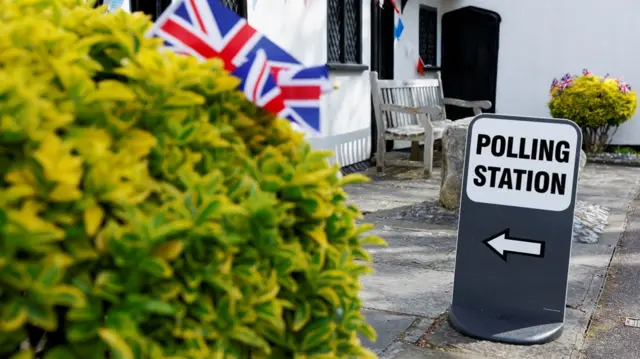 A polling station sign beside a hedge and some union flags