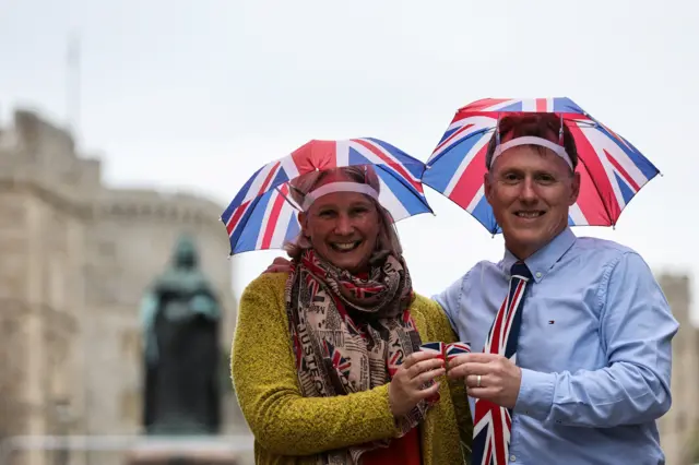 A couple wearing union flag umbrella hats toast each other outside Windsor Castle