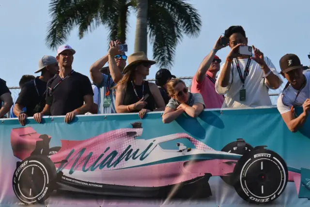 Fans lean on a banner of a pink F1 car with 'Miami' written on it