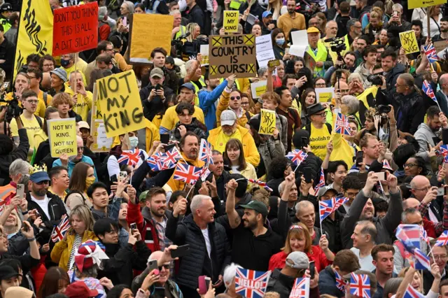 Crowds of people hold up anti-monarchy signs in front of royal fans waving union jacks