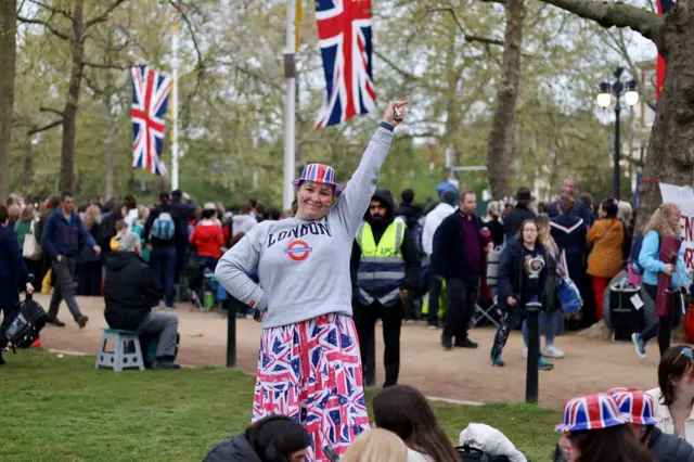 A woman in a London tube T-shirt and union jack trousers smiles and points on The Mall