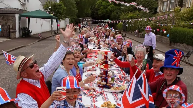 . Alt text: People sit at a table for a meal in a street, in a photo filled with union flags