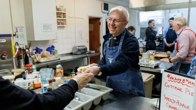 Justin Welby wears a denim apron with the words "Bon Jovi" on it, and serves cottage pie during a volunteering event