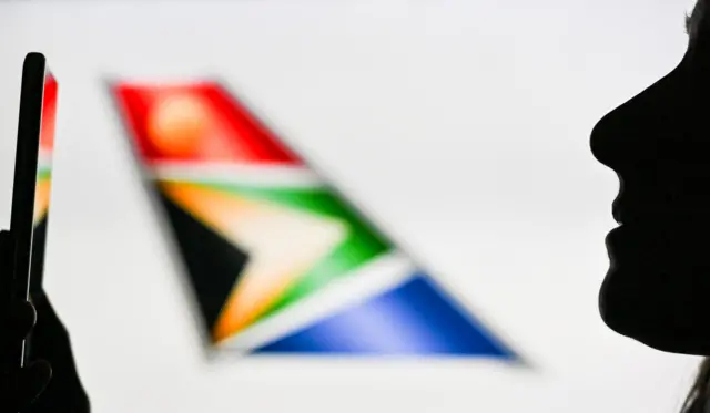 An image of a woman holding a cell phone in front of the South African Airways logo