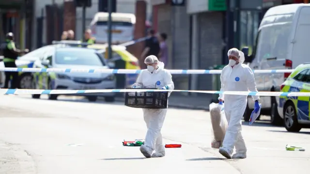 Two forensics officers, wearing white overalls, carry some equipment