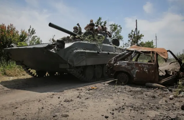 Ukrainian service members ride an infantry fighting vehicle next to a destroyed car