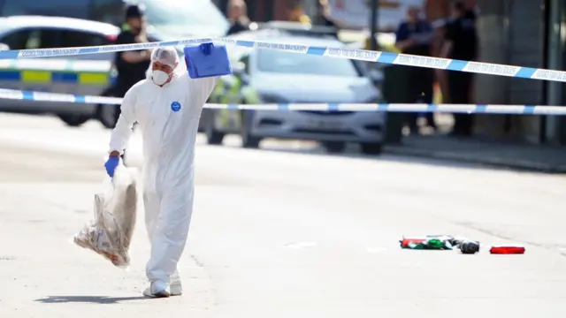 A police forensics officer, wearing a white overall, lifts up a police cordon to walk underneath it