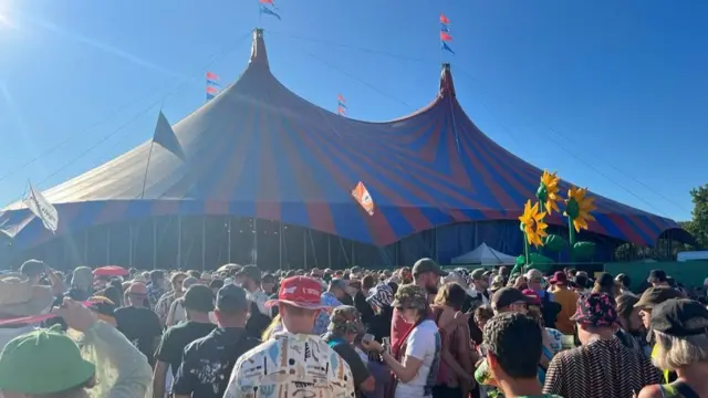 An exterior view of the tent in which Rick Astley is playing at Glastonbury. Large numbers of people are seen spilling out of the tent.