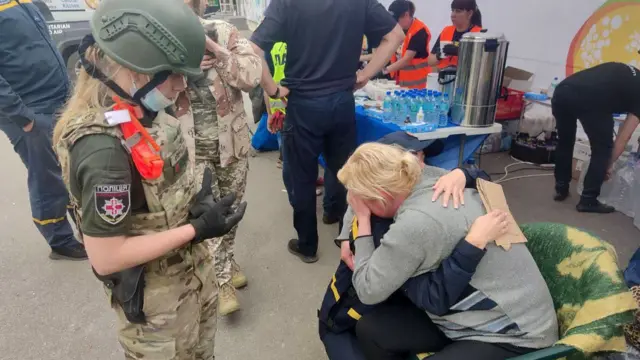 A Ukrainian rescue worker looks on as two people grieve in front of her