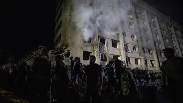 Rescuers work in the foreground while smoke billows from a building in the background