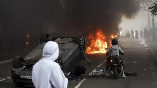 An overturned car, with a protester on a moped next to it and a burning car in front of them