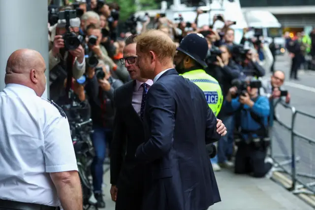 Prince Harry enters court escorted by a man in a suit, in front of gathered photographers