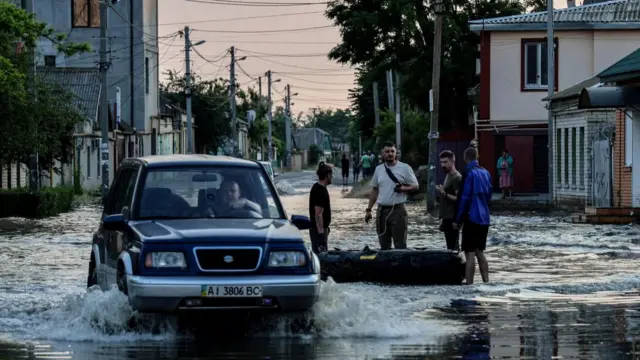 A car makes its way past people standing next to an inflatable boat, in a flooded street of Kherson