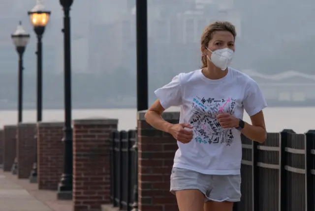 A woman wears a face mask as smoke continues to shroud the sun as it rises behind the skyline of Manhattan in New York City on June 7, 2023, as seen from Weehawken, New Jersey.