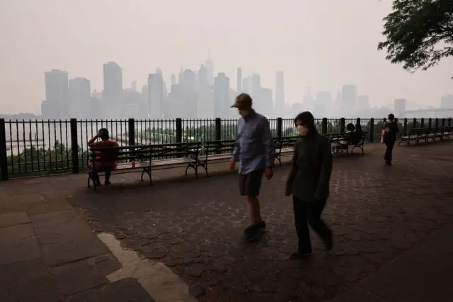 People walk along the Brooklyn Promenade as a reddish haze enshrouds the Manhattan skyline