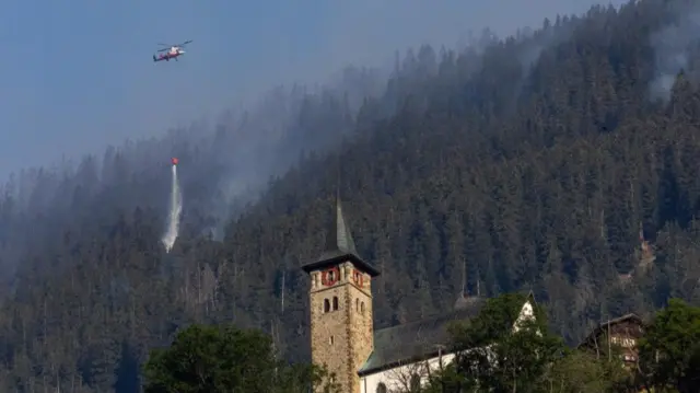 A helicopter carries water to a wildfire on the flank of a mountain in Bitsch