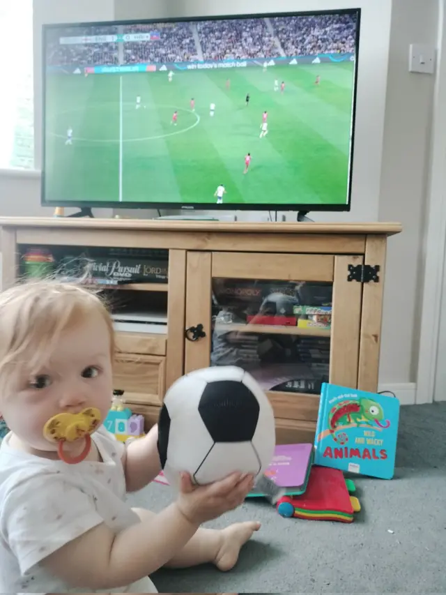 A young England fan watches the match on tv at home with a ball in her hands.