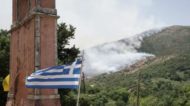 A Greek flag flies in the foreground, as smokes rises from a hillside near the village of Palia Perithia, on the island of Corfu, Greece