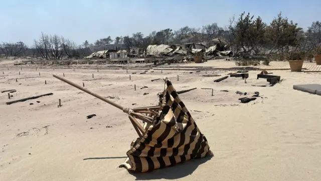 A charred and blown over parasol on the beach, with burned out huts in the background