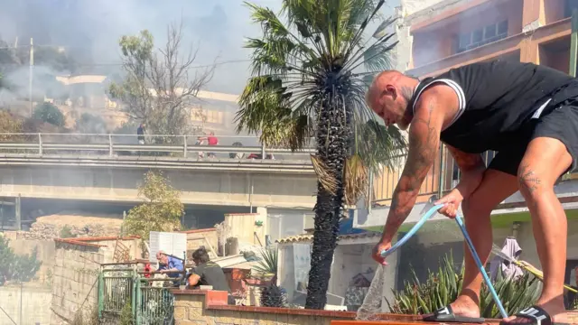 A man stands on a brick wall with a hose and sprays flames