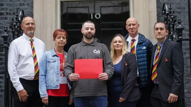 Four men and two women stand outside Number 10 Downing Street, one man is holding a large a4 sized red envelope.