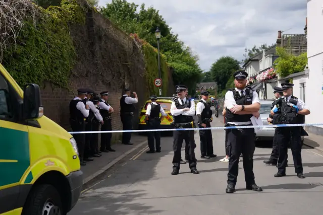 Police officers stand on a taped off Camp Road, an ambulance is seen in in the foreground