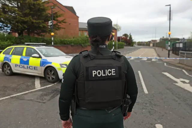 A PSNI officers stands with their back to the camera at the end of a closed off road with a police car in the distance