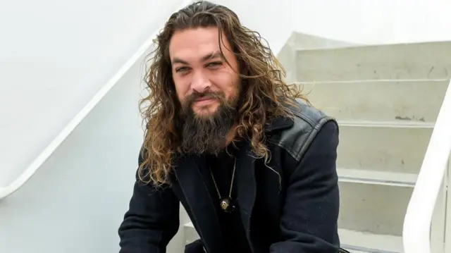 Jason Momoa sits on a staircase, smiling at the camera