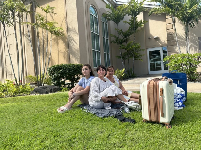 A grandmother, mother and daughter sitting on a manicured lawn with their luggage in front of a villa