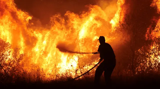 A silhouetted firefighter battles massive flames in centrak Greece