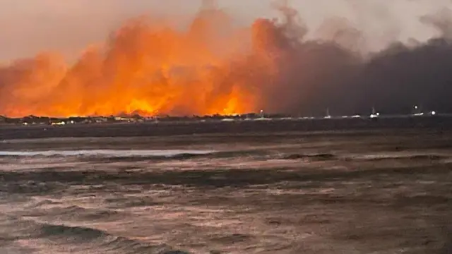 A distant view of flames in Lahaina, with waves in the foreground