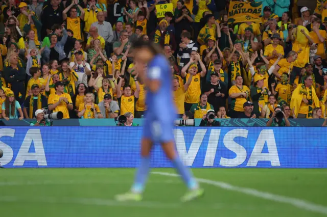 Matildas fans roar on their side with a French player walking past in the foreground.