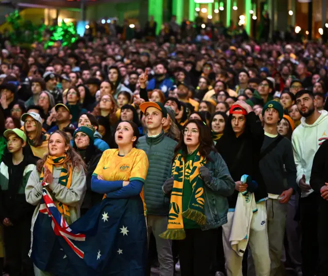 Australia fans stand watching the big screen in a fan park.