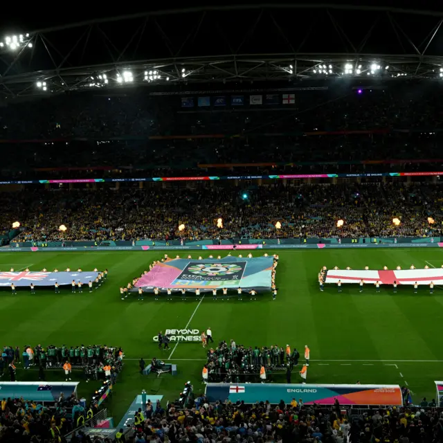 Both team flags flank the competition logo on the pitch.