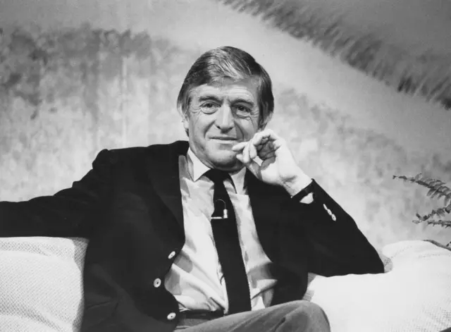 A black and white photo of sir Parkinson sitting on a couch on the set of a television show in 1985.