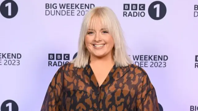 Radio 1 DJ Katie Thistleton. Katie, a white, blonde woman in her 30s, is pictured at Radio 1's Big Weekend in Dundee. Katie wears a sheer orange and brown animal print blouse and smiles at the camera.