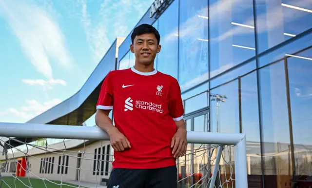 Wataru Endo leans on a goalpost to pose in his new Liverpool shirt.