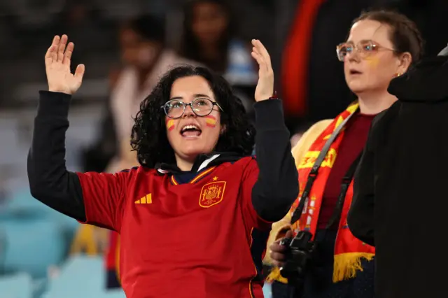 A Spain fan chants with her arms in the air while wearing the shirt and having flags painted on her face.