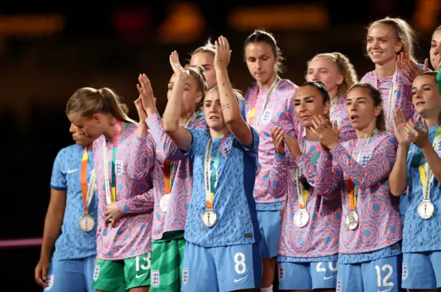 England players stand on the podium with their runners up medals clapping the crowd.