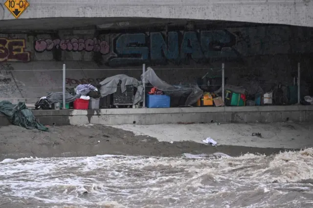 Tents and belongings of homeless people are seen near the rushing water of the Los Angeles River