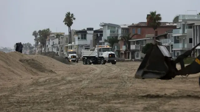 Trucks drive along a beach, beside enormous constructed sand walls in front of lavish houses