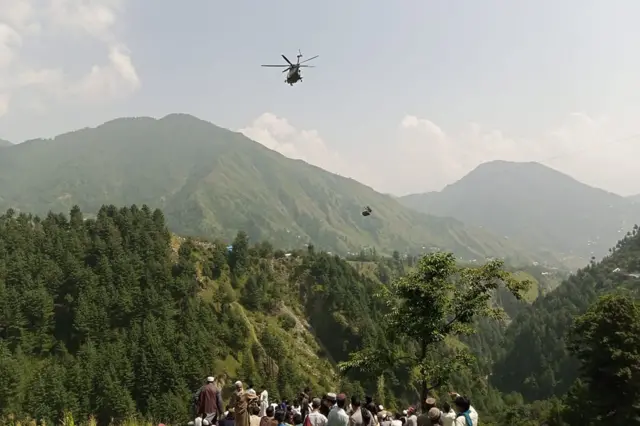 People on a mountainside watch as an army helicopter hovers over a suspended chairlift in the mountains